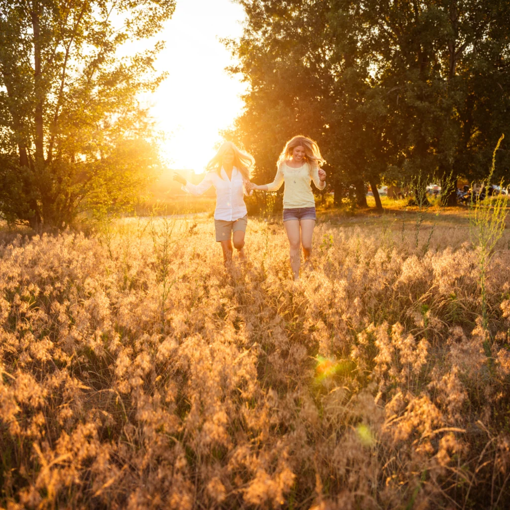 Two women running hand in hand through sunny field of tall grass.