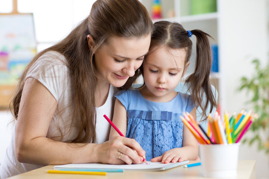 Mother helping young daughter write in notebook with colored pencils.