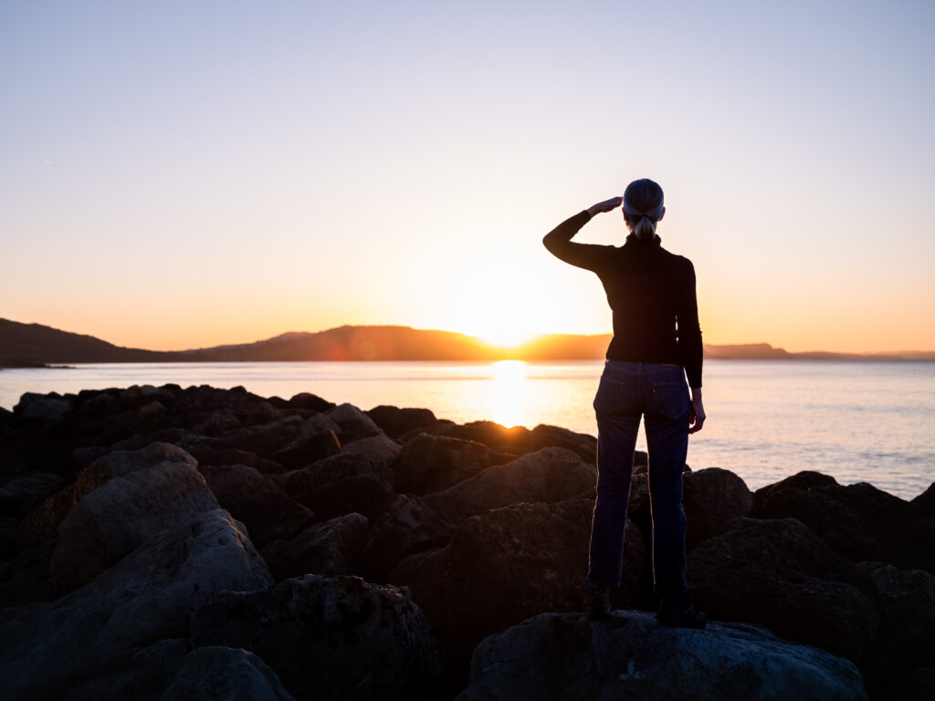Person standing on rocks saluting sunset over calm water.